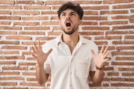 Foto de Hombre árabe con barba de pie sobre ladrillos fondo de la pared loco y loco gritando y gritando con expresión agresiva y los brazos levantados. concepto de frustración. - Imagen libre de derechos