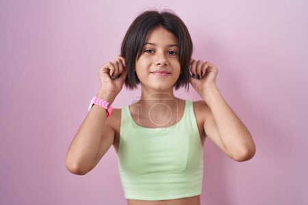 Photo for Young girl standing over pink background smiling pulling ears with fingers, funny gesture. audition problem - Royalty Free Image