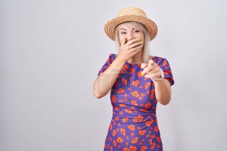 Téléchargez les photos : Jeune femme caucasienne portant des fleurs robe et chapeau d'été se moquant de vous, pointant du doigt la caméra avec la main sur la bouche, expression de honte - en image libre de droit