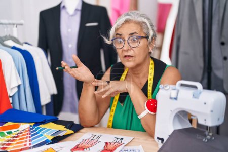 Téléchargez les photos : Middle age grey-haired woman tailor sitting on table speaking at tailor shop - en image libre de droit