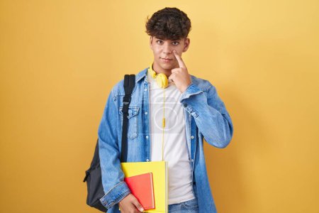 Téléchargez les photos : Adolescent hispanique portant un sac à dos d'étudiant et tenant des livres pointant vers l'œil vous regardant geste, expression suspecte - en image libre de droit