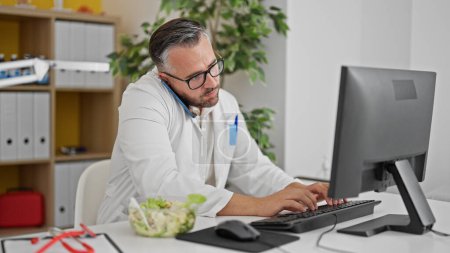 Photo for Grey-haired man doctor talking on smartphone using computer at the clinic - Royalty Free Image