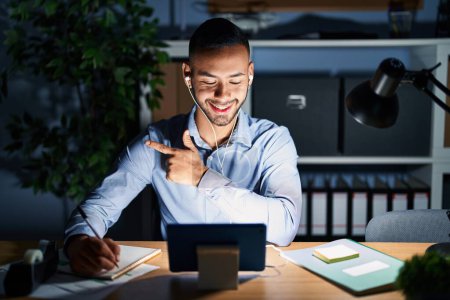 Téléchargez les photos : Jeune homme hispanique travaillant au bureau la nuit gai avec un sourire sur le visage pointant avec la main et le doigt sur le côté avec une expression heureuse et naturelle - en image libre de droit