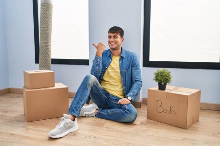 Photo for Young hispanic man sitting on the floor at new home smiling with happy face looking and pointing to the side with thumb up. - Royalty Free Image