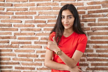 Téléchargez les photos : Jeune adolescente debout sur le mur de briques pointant avec le doigt de la main sur le côté montrant la publicité, visage sérieux et calme - en image libre de droit