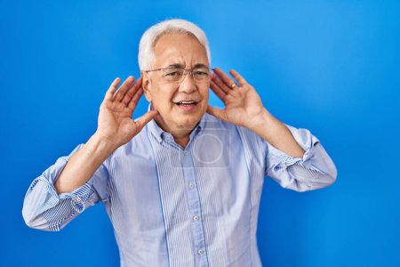 Foto de Hombre mayor hispano usando anteojos tratando de escuchar el gesto de ambas manos en la oreja, curioso por chismes. problema auditivo, sordo - Imagen libre de derechos