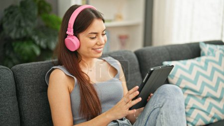 Téléchargez les photos : Jeune belle femme hispanique souriant confiant en utilisant touchpad et écouteurs à la maison - en image libre de droit