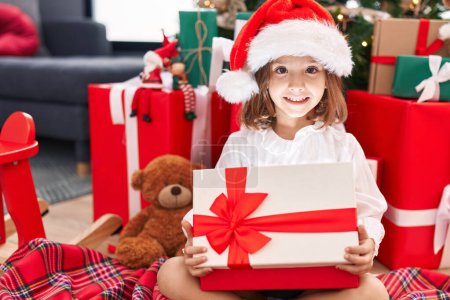 Photo for Adorable hispanic girl unpacking gift sitting by christmas tree at home - Royalty Free Image