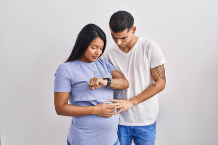 Téléchargez les photos : Jeune couple hispanique s'attendant à un bébé debout sur fond de vérification de l'heure sur la montre-bracelet, détendu et confiant - en image libre de droit