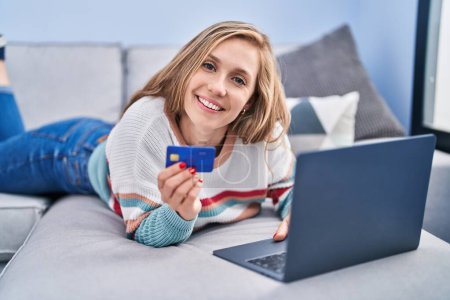 Photo for Young blonde woman using laptop and credit card lying on sofa at home - Royalty Free Image