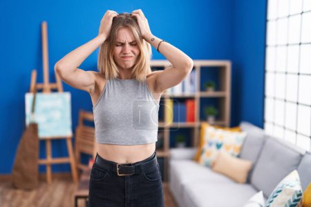Photo for Blonde caucasian woman standing at living room suffering from headache desperate and stressed because pain and migraine. hands on head. - Royalty Free Image