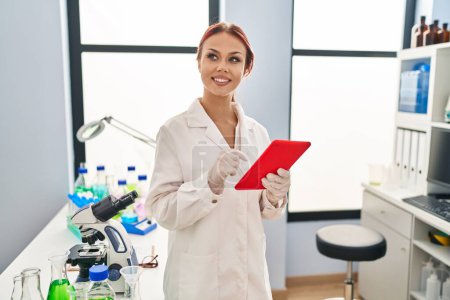 Téléchargez les photos : Jeune femme caucasienne scientifique souriant confiant en utilisant le pavé tactile au laboratoire - en image libre de droit
