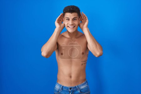 Téléchargez les photos : Jeune homme hispanique debout torse nu sur fond bleu essayant d'entendre les deux mains sur le geste de l'oreille, curieux de commérages. problème auditif, sourd - en image libre de droit