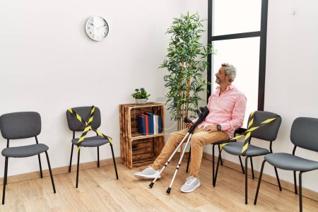 Téléchargez les photos : Homme aux cheveux gris d'âge moyen blessé assis sur une chaise tenant des béquilles dans la salle d'attente de la clinique - en image libre de droit