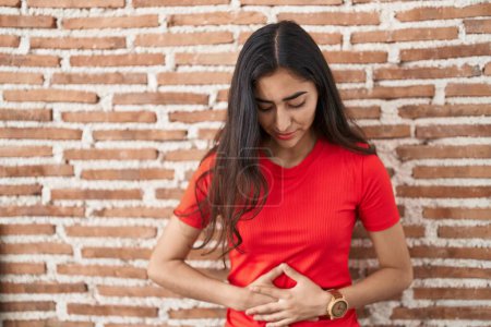 Photo for Young teenager girl standing over bricks wall with hand on stomach because indigestion, painful illness feeling unwell. ache concept. - Royalty Free Image
