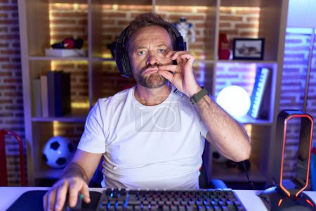 Téléchargez les photos : Homme d'âge moyen avec barbe jouant à des jeux vidéo portant casque bouche et lèvres fermées comme zip avec les doigts. secret et silencieux, tabou parlant - en image libre de droit