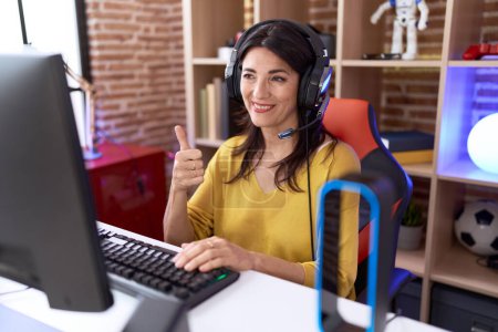 Foto de Mujer hispana de mediana edad jugando videojuegos usando auriculares sonriendo feliz y positivo, pulgar hacia arriba haciendo excelente y signo de aprobación - Imagen libre de derechos