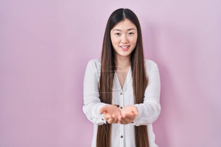 Foto de Mujer joven china de pie sobre fondo rosa sonriendo con las palmas de las manos juntas recibiendo o dando gesto. retención y protección - Imagen libre de derechos