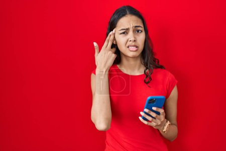 Foto de Mujer brasileña joven usando teléfono inteligente sobre fondo rojo disparando y matándose señalando la mano y los dedos a la cabeza como arma, gesto suicida. - Imagen libre de derechos