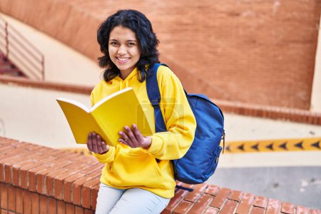 Foto de Joven estudiante latina sonriendo confiada leyendo libro en la calle - Imagen libre de derechos