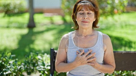 Foto de Mujer de mediana edad respirando con las manos en el pecho sentado en el banco en el parque - Imagen libre de derechos