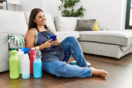 Photo for Young beautiful hispanic woman using smartphone sitting on floor with clean products at home - Royalty Free Image