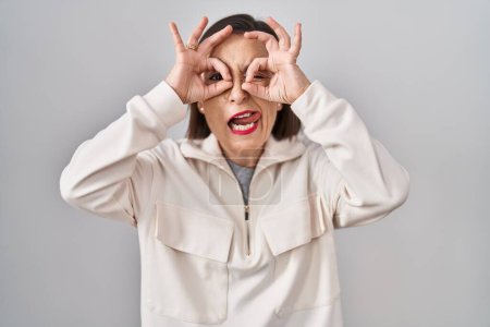 Foto de Mujer hispana de mediana edad de pie sobre un fondo aislado haciendo un buen gesto como binoculares sacando la lengua, ojos mirando a través de los dedos. expresión loca. - Imagen libre de derechos