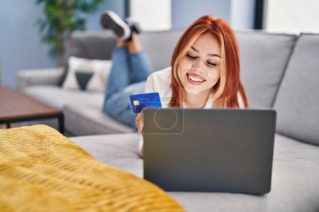 Téléchargez les photos : Jeune femme caucasienne utilisant ordinateur portable et carte de crédit couché sur le canapé à la maison - en image libre de droit