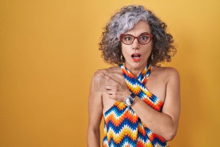 Foto de Mujer de mediana edad con el pelo gris de pie sobre el fondo amarillo sorprendido señalando con el dedo hacia el lado, boca abierta expresión asombrada. - Imagen libre de derechos
