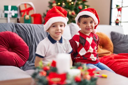 Foto de Dos niños sonriendo confiados sentados en el sofá junto al árbol de Navidad en casa - Imagen libre de derechos