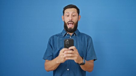 Téléchargez les photos : Jeune homme hispanique utilisant un smartphone avec expression surprise sur fond bleu isolé - en image libre de droit