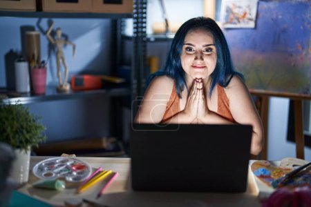 Téléchargez les photos : Jeune fille moderne avec les cheveux bleus assis au studio d'art avec ordinateur portable la nuit priant avec les mains ensemble demandant pardon souriant confiant. - en image libre de droit