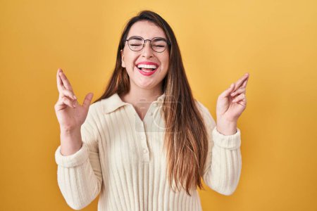Téléchargez les photos : Jeune femme hispanique debout sur fond jaune geste doigt croisé souriant avec espoir et les yeux fermés. chance et concept superstitieux. - en image libre de droit