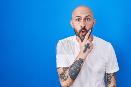 Téléchargez les photos : Homme hispanique avec des tatouages debout sur fond bleu regardant fasciné par l'incrédulité, la surprise et l'expression étonnante avec les mains sur le menton - en image libre de droit