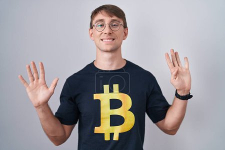 Foto de Hombre rubio caucásico con camiseta bitcoin mostrando y apuntando hacia arriba con los dedos número nueve mientras sonríe confiado y feliz. - Imagen libre de derechos