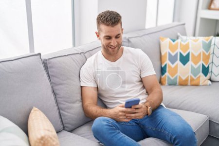 Foto de Young caucasian man using smartphone sitting on sofa at home - Imagen libre de derechos