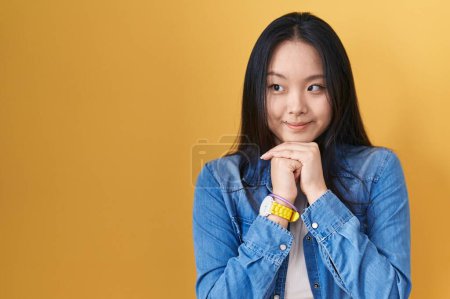 Foto de Joven mujer asiática de pie sobre fondo amarillo riendo nervioso y excitado con las manos en la barbilla mirando a un lado - Imagen libre de derechos