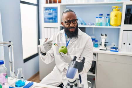 Foto de Hombre afroamericano que trabaja en el laboratorio científico con expresión de manzana despistada y confusa. concepto de duda. - Imagen libre de derechos