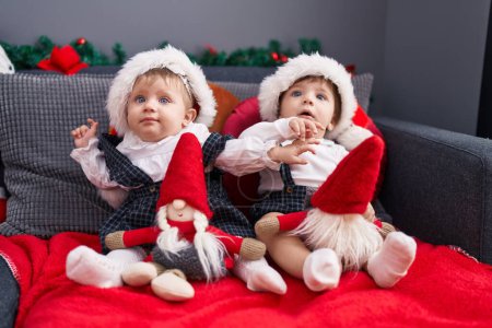 Photo for Two adorable babies sitting on sofa playing with santa claus doll at home - Royalty Free Image