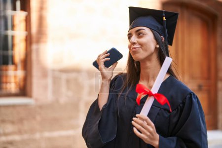 Photo for Young hispanic woman wearing graduated uniform holding diploma hearing audio message at university - Royalty Free Image