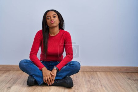 Foto de Joven afroamericano con trenzas sentado en el suelo en casa mirando a la cámara soplando un beso en el aire siendo encantador y sexy. expresión de amor. - Imagen libre de derechos