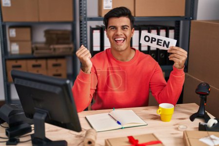 Photo for Hispanic man working at small business ecommerce holding open banner screaming proud, celebrating victory and success very excited with raised arms - Royalty Free Image