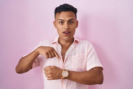 Téléchargez les photos : Jeune homme hispanique debout sur fond rose à la hâte pointant pour regarder le temps, l'impatience, bouleversé et en colère pour retard de délai - en image libre de droit