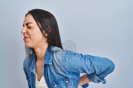 Téléchargez les photos : Femme hispanique debout sur fond bleu souffrant de maux de dos, toucher le dos avec la main, douleurs musculaires - en image libre de droit