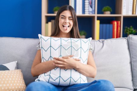 Téléchargez les photos : Femme hispanique étreignant oreiller assis sur le canapé langue collante heureux avec une expression drôle. - en image libre de droit