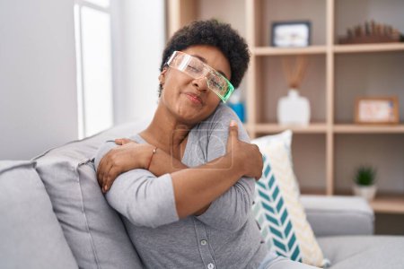 Téléchargez les photos : Jeune femme afro-américaine portant des lunettes de réalité virtuelle assise sur le canapé se serrant dans ses bras heureux et positifs, souriant confiant. amour de soi et soins de soi - en image libre de droit