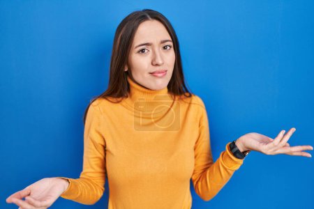 Photo for Young brunette woman standing over blue background clueless and confused with open arms, no idea concept. - Royalty Free Image