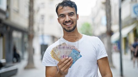 Foto de Joven hispano sonriendo confiado sosteniendo pesos mexicanos en la calle - Imagen libre de derechos