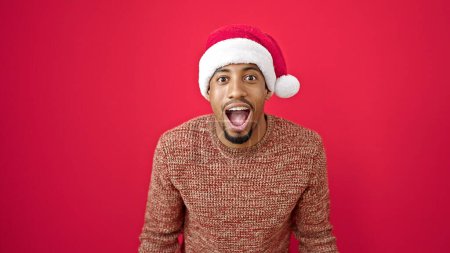 Téléchargez les photos : Afro-Américain expression surprise portant chapeau de Noël sur fond rouge isolé - en image libre de droit