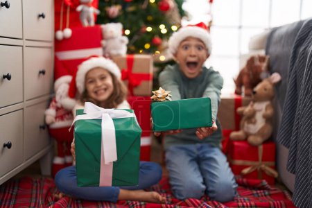 Téléchargez les photos : Deux enfants tenant un cadeau assis sur le sol près de l'arbre de Noël à la maison - en image libre de droit